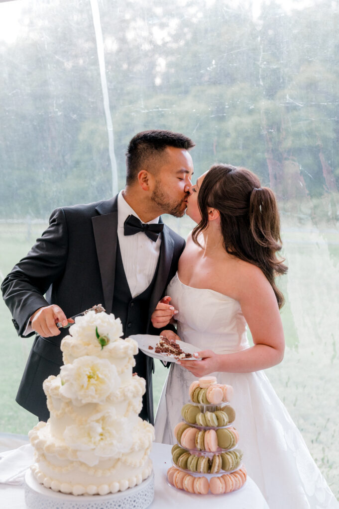 Bride and groom share a kiss during their cake cutting moment, with white tiered wedding cake and macaron tower on display.