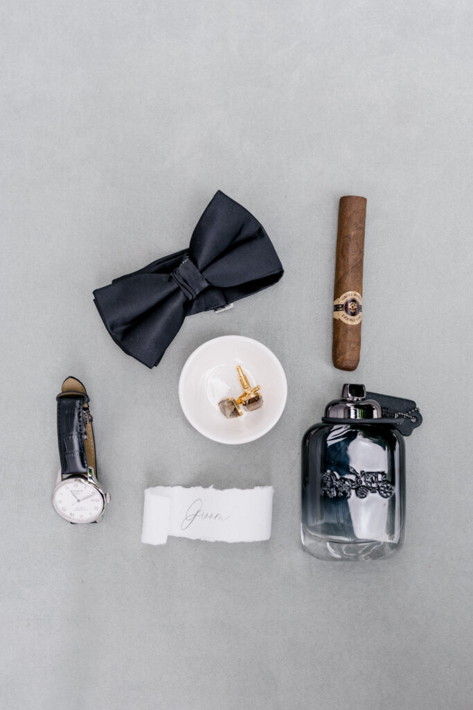 Fine art groom details with black tuxedo accessories, cologne, and boutonniere on a gray linen background