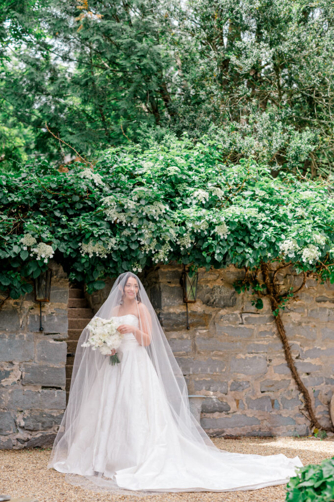 Bride in strapless white gown and long veil holding white bouquet, standing against stone wall and lush green flowering vines at a Connecticut wedding venue.