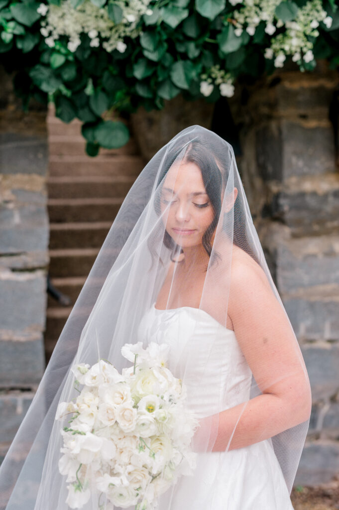 Close-up portrait of the bride under a veil holding a white garden-style bouquet.