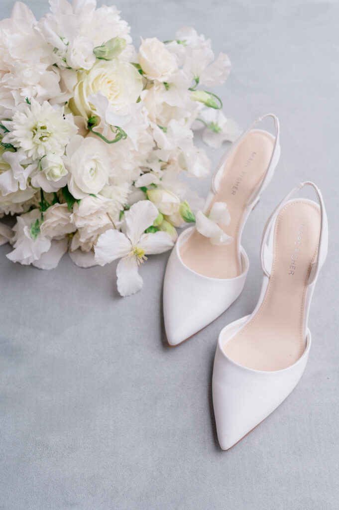 Fine art wedding flatlay of white pointed heels, lush bouquet, and bride’s accessories on soft linen