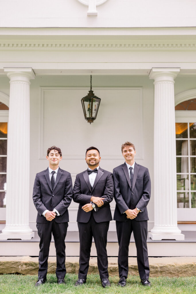 Groom and groomsmen in black tuxedos posing in front of white columns at Smith Farm Gardens.