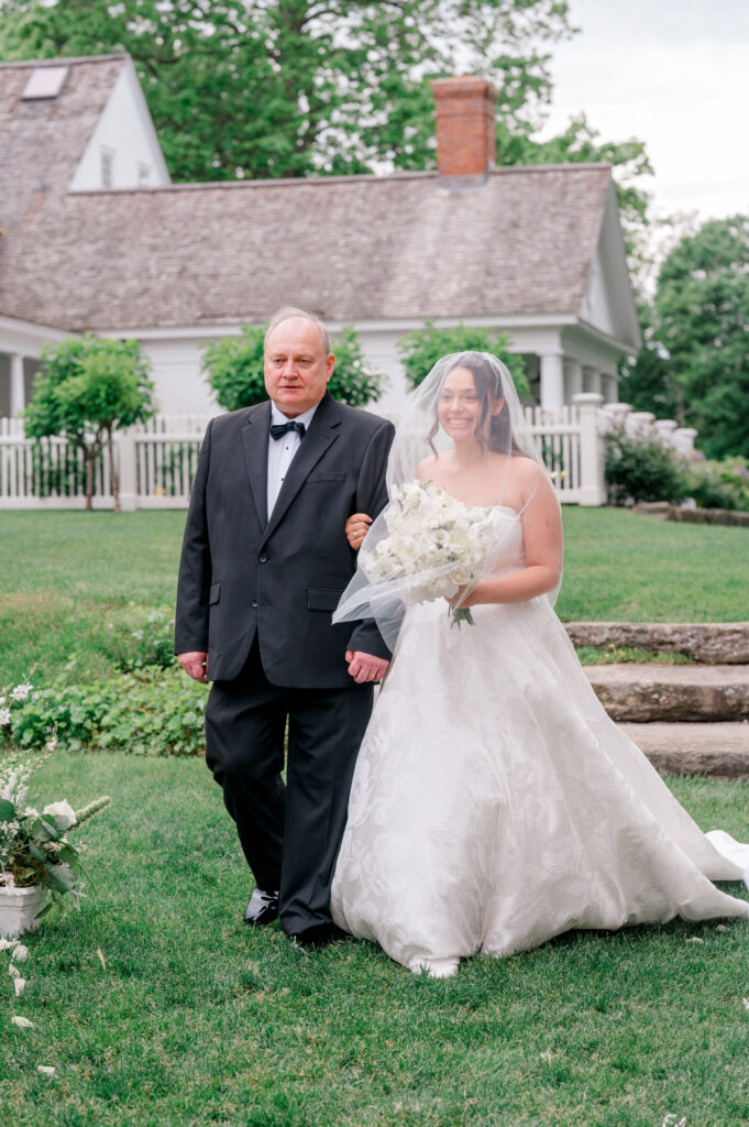 The bride walks down the aisle with her father, smiling under her veil at Smith Farm Gardens