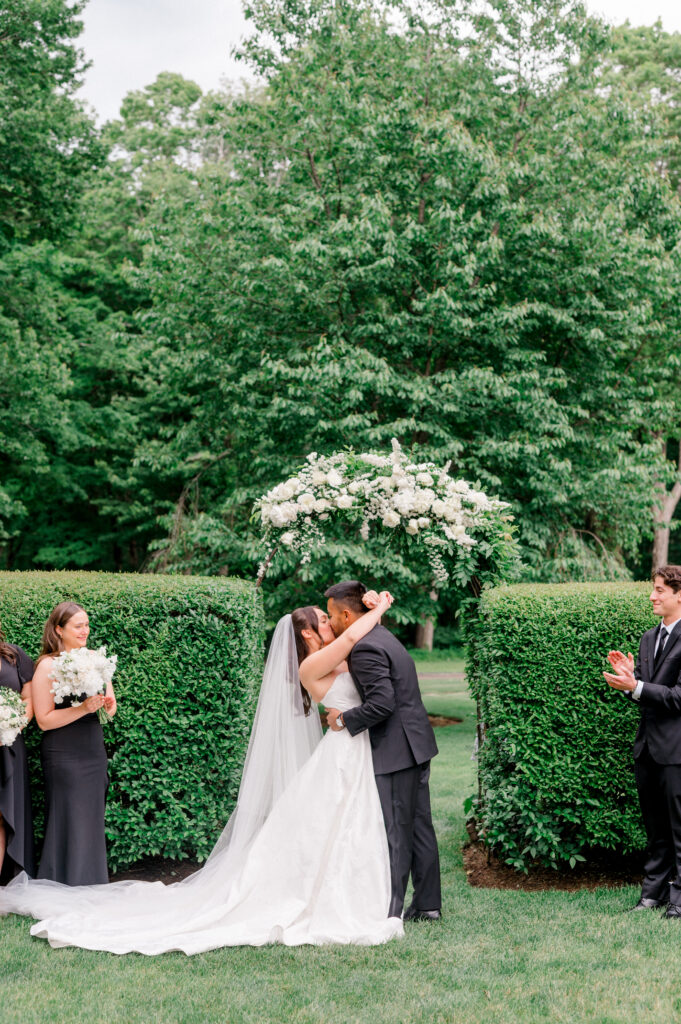 Emily and Patrick kiss at the altar beneath a lush white floral arch during their Smith Farm Gardens ceremony