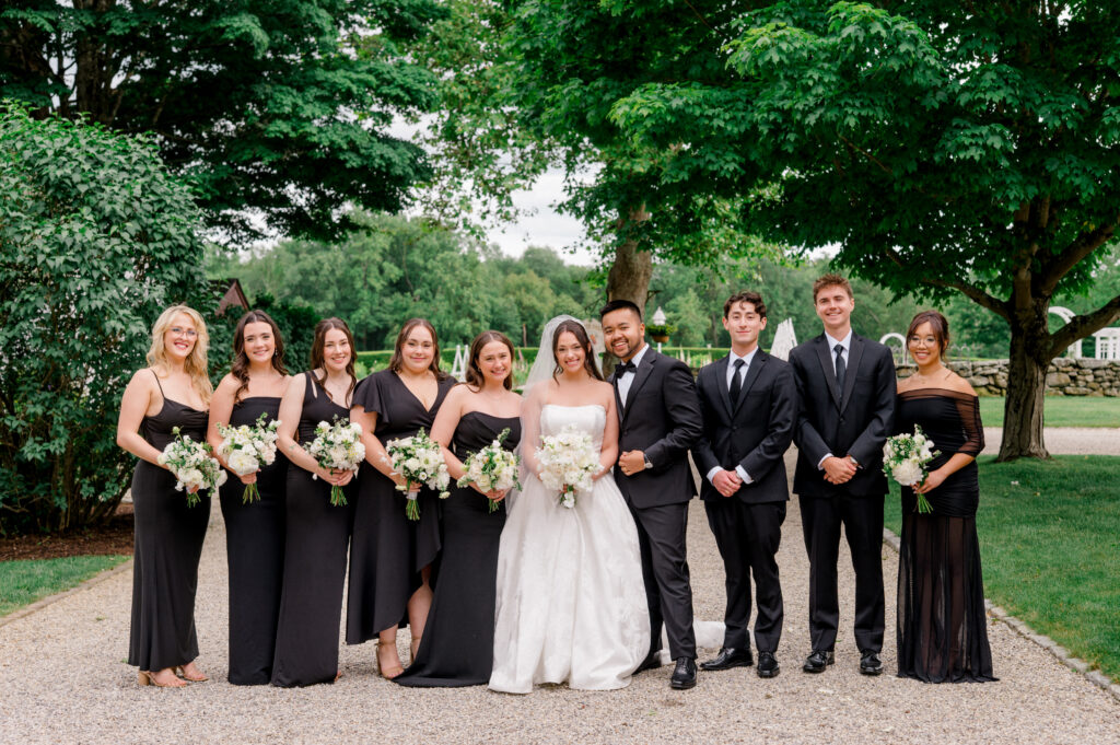 Wedding party group portrait at Smith Farm Gardens with the bride in a strapless gown and bridesmaids in black dresses holding white bouquets.
