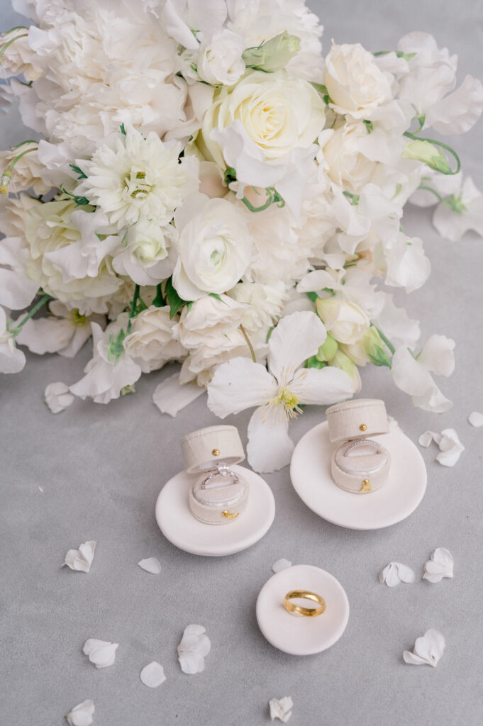 Wedding rings displayed in velvet ring boxes next to a white floral bouquet, styled on a soft gray background by Jennifer Nichole Photography.