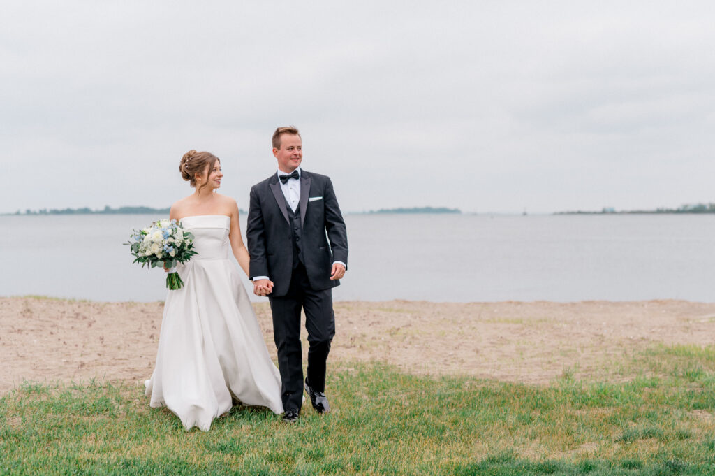 Bride and groom walk hand in hand along the sandy shore during their waterfront wedding at The Point at Norwalk Cove, bride in a modern strapless gown holding a bouquet of white and blue flowers. Photographed by Jennifer Nichole Photography, Connecticut wedding photographer known for heirloom and coastal wedding photography.