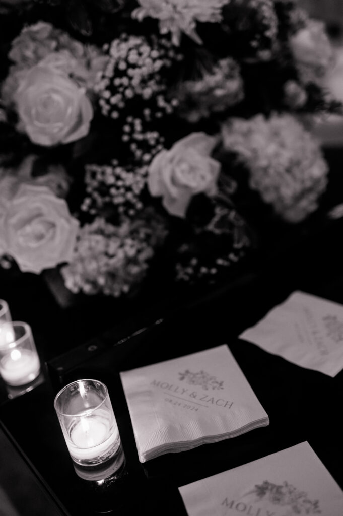 Black-and-white detail photo of custom wedding cocktail napkins surrounded by candles and florals, illustrating reception details in a Connecticut wedding photography timeline captured by Jennifer Nichole.