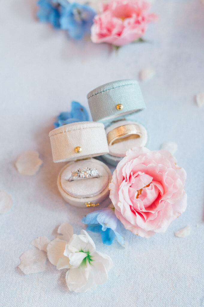 Close-up of cream and blue velvet ring boxes with diamond and gold wedding bands beside pink, blue, and white florals.