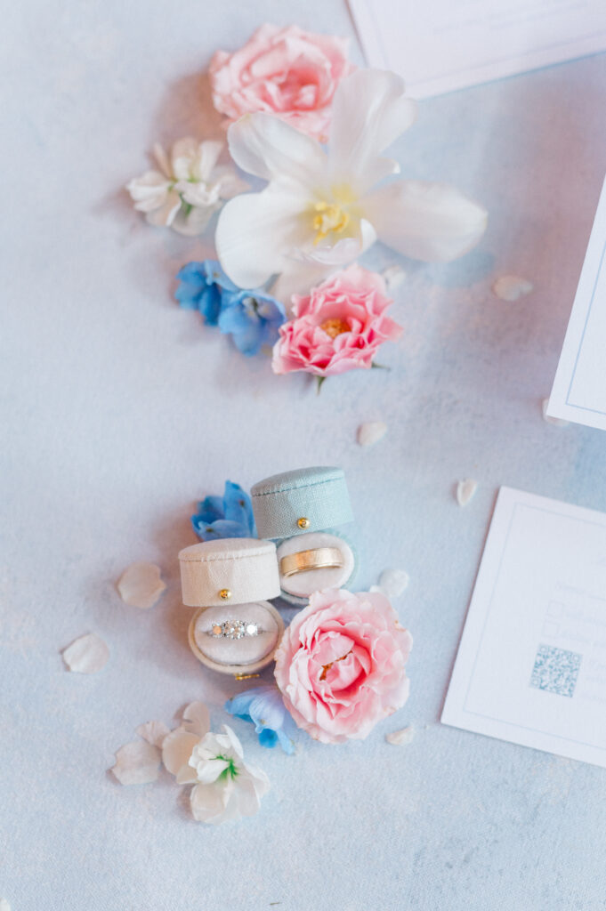 Velvet ring boxes with gold and diamond bands styled with pastel florals and blue invitation accents for romantic wedding flatlay photography.