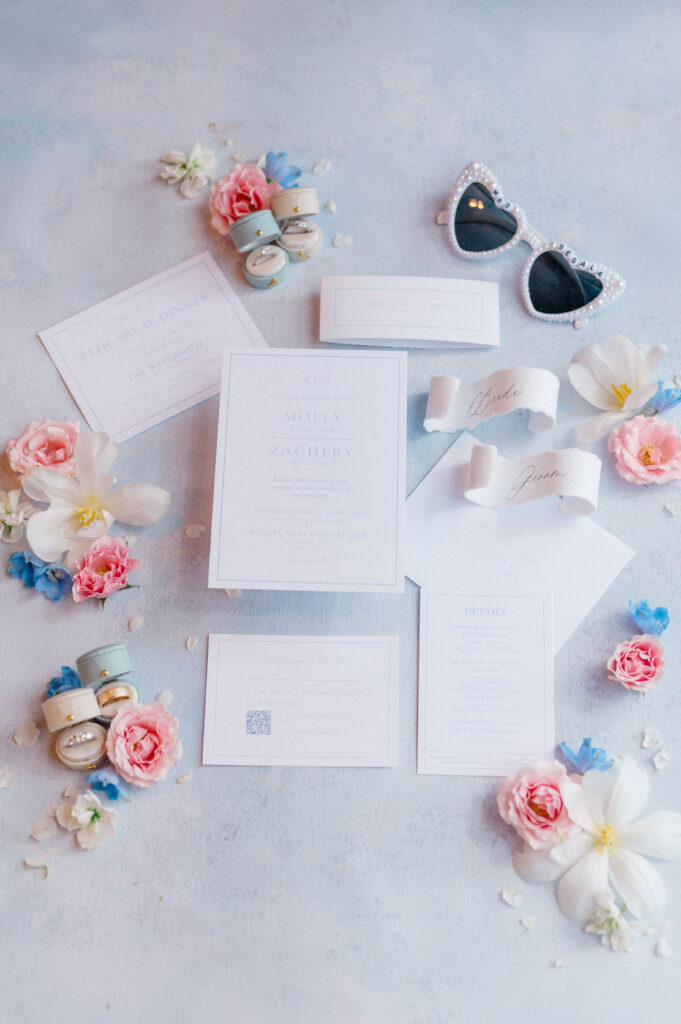 Full bridal flatlay with invitation suite, pearl sunglasses, vow ribbons, and ring boxes surrounded by pink, white, and blue floral accents.
