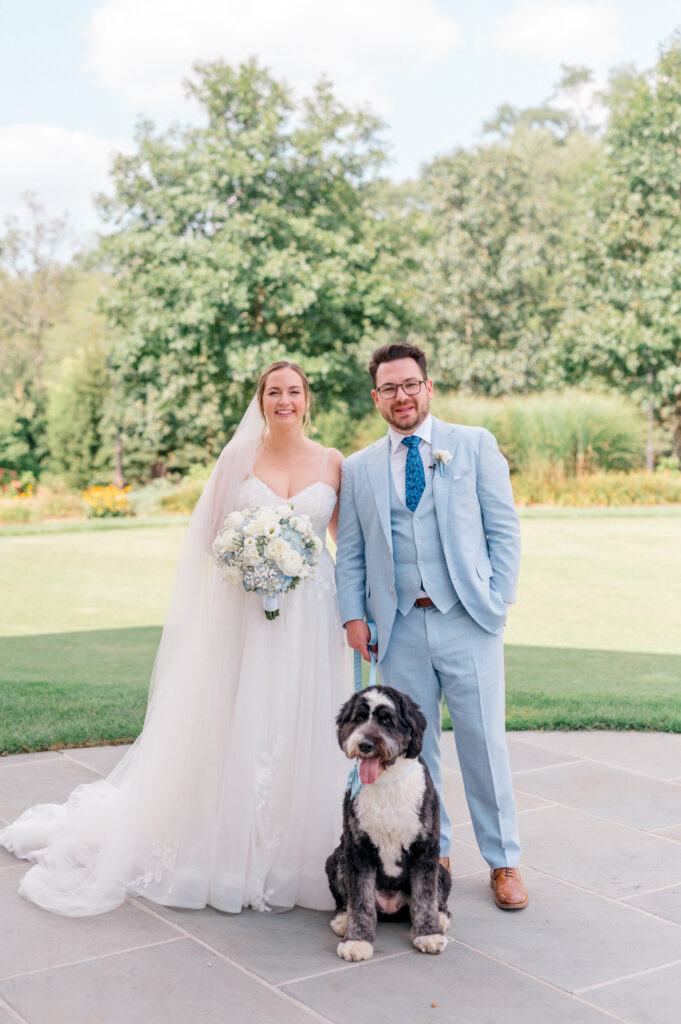 Bride in tulle gown with sweetheart neckline and groom in light blue suit posing with black and white dog on stone terrace, with green trees and garden backdrop.