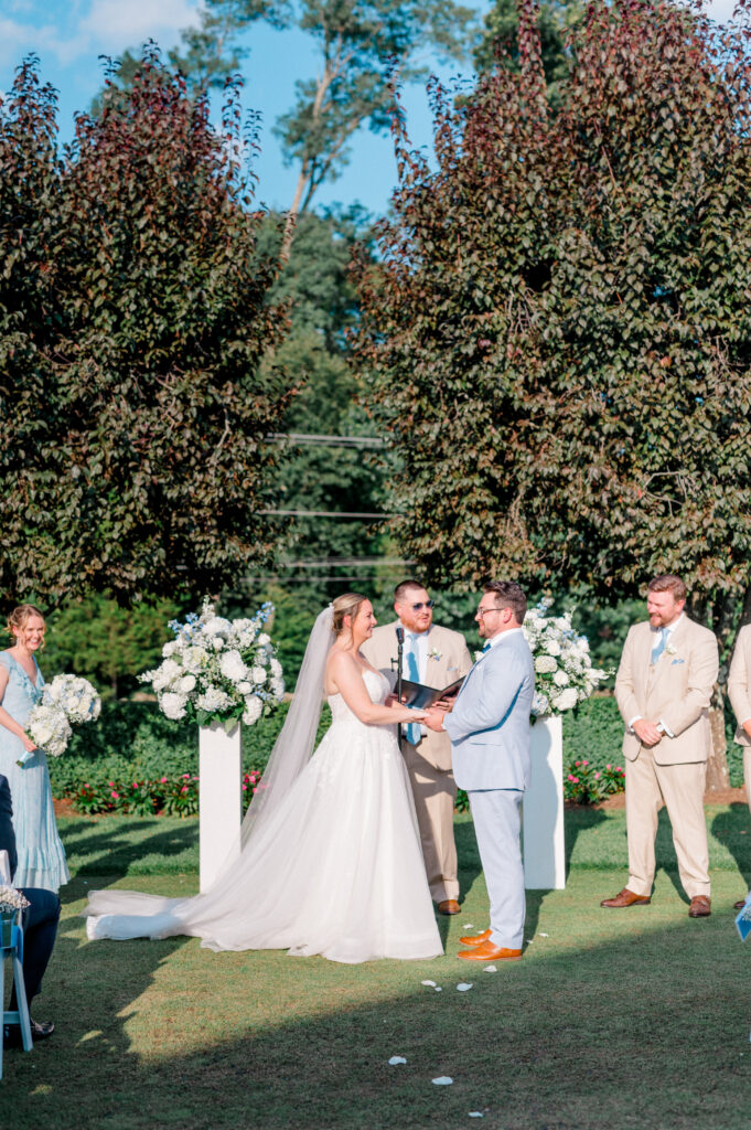 Bride and groom exchange vows outdoors under blue sky, surrounded by white floral arrangements and bridal party, captured by Connecticut wedding photographer Jennifer Nichole.