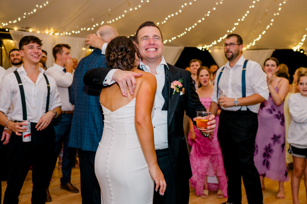 Groom hugging bride under twinkling lights while guests cheer on the dance floor, part of evening celebrations in a Connecticut wedding photography timeline photographed by Jennifer Nichole & Co.