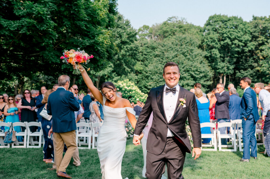 Joyful bride in off-shoulder gown raises vibrant bouquet while walking hand-in-hand with groom down the aisle, celebrating during Connecticut wedding ceremony at Smith Farm Gardens photographed by Jennifer Nichole.