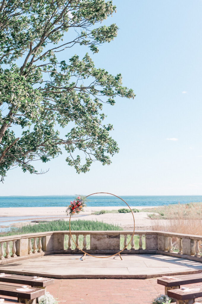 Oceanfront wedding ceremony setup featuring circular floral arch, beach view, and clear blue sky, styled for elegant Connecticut weddings at Eolia Mansion at Harkness Park by Jennifer Nichole Photography.