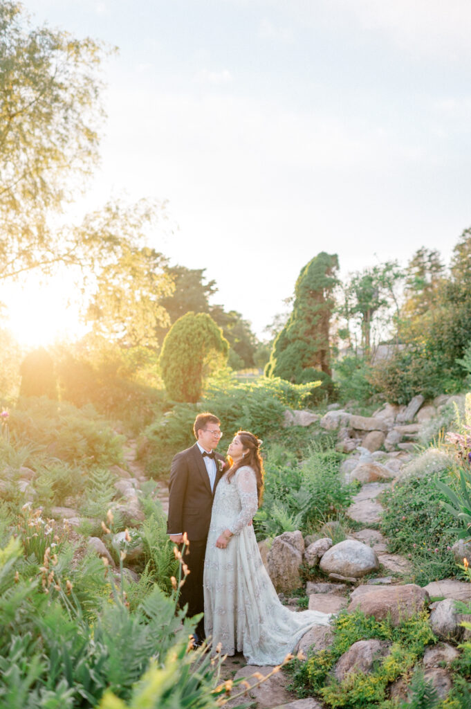 Bride and groom standing in sunlit garden surrounded by lush greenery and rocks, sharing a romantic moment during golden hour portraits at Eolia Mansion.