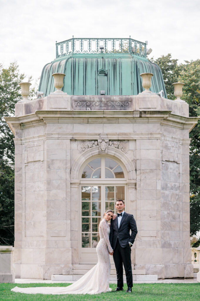 Bride in long-sleeve lace gown and groom in black tuxedo standing hand in hand in front of historic stone pavilion with green copper dome at The Elms Mansion in Newport Rhode Island.