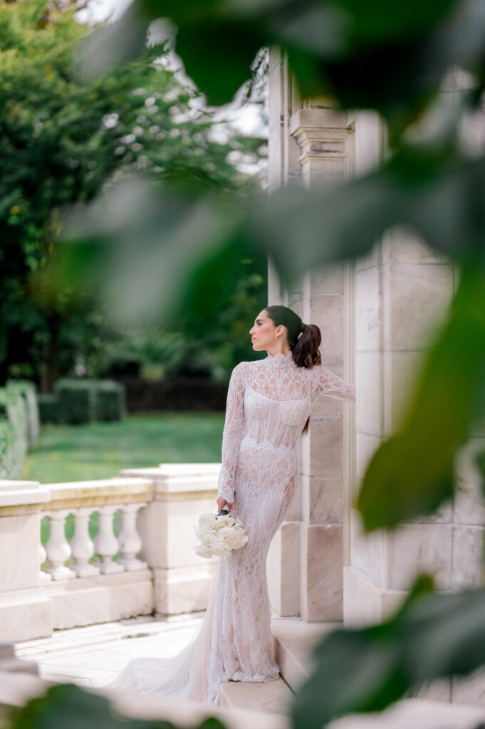 Bride in high-neck, long-sleeve lace wedding dress holding white bouquet, standing elegantly beside marble building exterior in soft natural light.